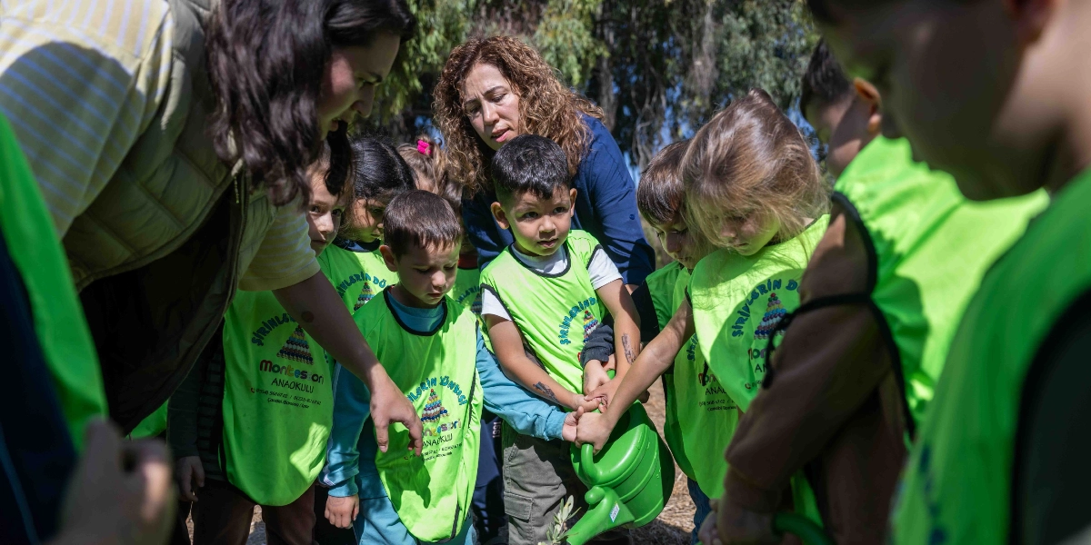 Üzerlerinde Şirinlerin Dünyası Montessori Anaokulu yazılı neon yeşil yelekler giymiş bir grup çocuk, öğretmenleri eşliğinde dışarıda bir fidanı suluyor. Çocuklar hep birlikte yeşil bir sulama kabını tutarak toprağa su döküyor, öğretmenleri ise onlara rehberlik ederek dikkatle izliyor.
