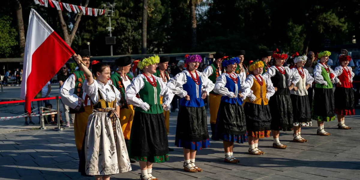 Geleneksel Polonya kıyafetleri giymiş bir grup dansçı, açık havada yan yana dizilmiş şekilde duruyor. Kadınlar beyaz gömlekler, renkli yelekler, çiçekli taçlar ve işlemeli etekler giyerken, erkekler yelek ve pantolonlardan oluşan yöresel kostümlerle onlara eşlik ediyor. Grubun en önündeki kadın, Polonya bayrağını havaya kaldırarak tutuyor. Arka planda ağaçlar ve izleyiciler görülüyor.