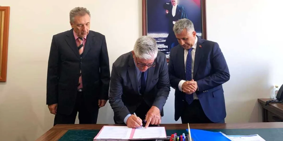 Takım elbiseli üç erkek, ofis ortamında bir masanın başında duruyor. Ortada oturan kişi bir belgeyi imzalarken, yanındaki iki kişi onu dikkatle izliyor. Arka planda duvarda Atatürk'ün bir portresi asılı.