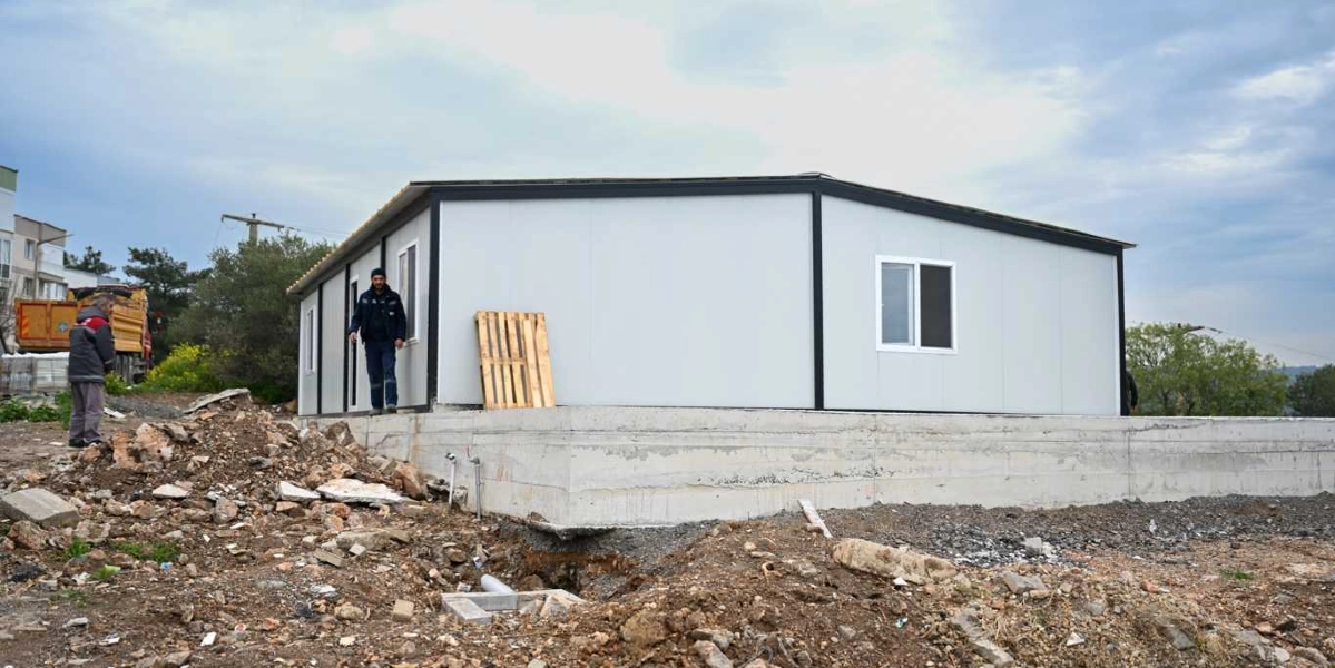 Beton bir temel üzerine inşa edilmiş, beyaz panellerden oluşan tek katlı prefabrik bir yapı. Yapının önünde bir işçi duruyor, sol tarafta ise inşaat alanında park etmiş sarı bir kamyon ve başka bir çalışan görülüyor. Zemin toprak ve molozlarla kaplı, gökyüzü ise bulutlu.