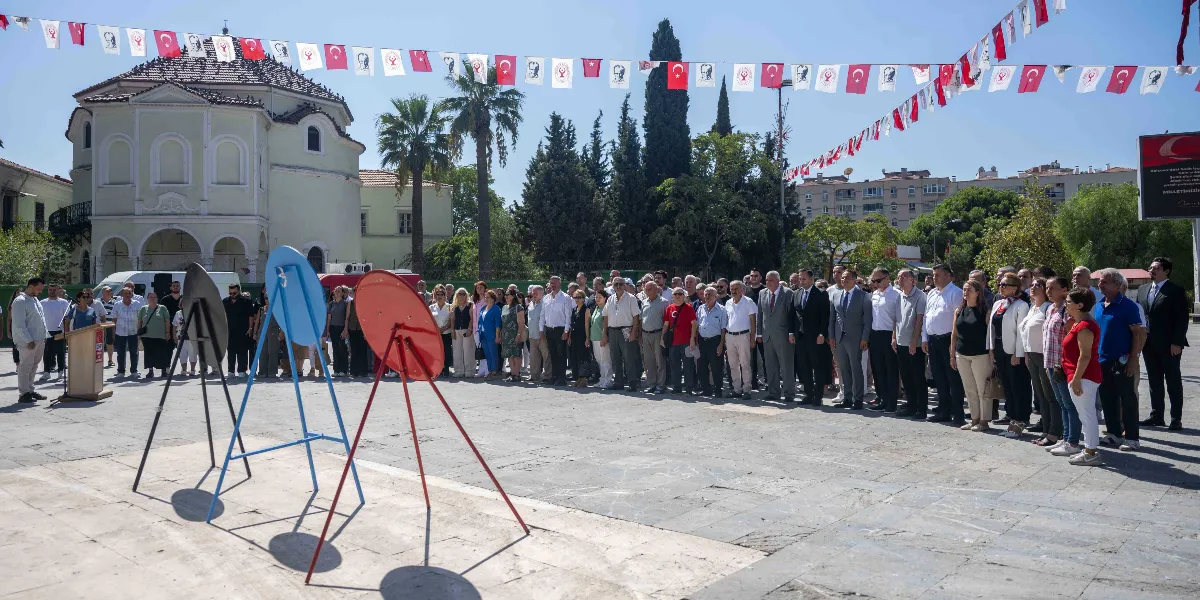 Bornova'da düzenlenen 9 Eylül kutlamalarında, meydanda toplanmış kalabalık bir grup insan, önlerinde duran üç adet boş çelenk standına doğru dönmüş şekilde duruyor. Meydanın üzerinde Türk bayrakları ve parti flamaları asılı. Arka planda tarihi bir yapı, palmiye ağaçları ve şehir binaları görülüyor. Sol tarafta bir kürsüde konuşma yapan bir kişi bulunuyor.