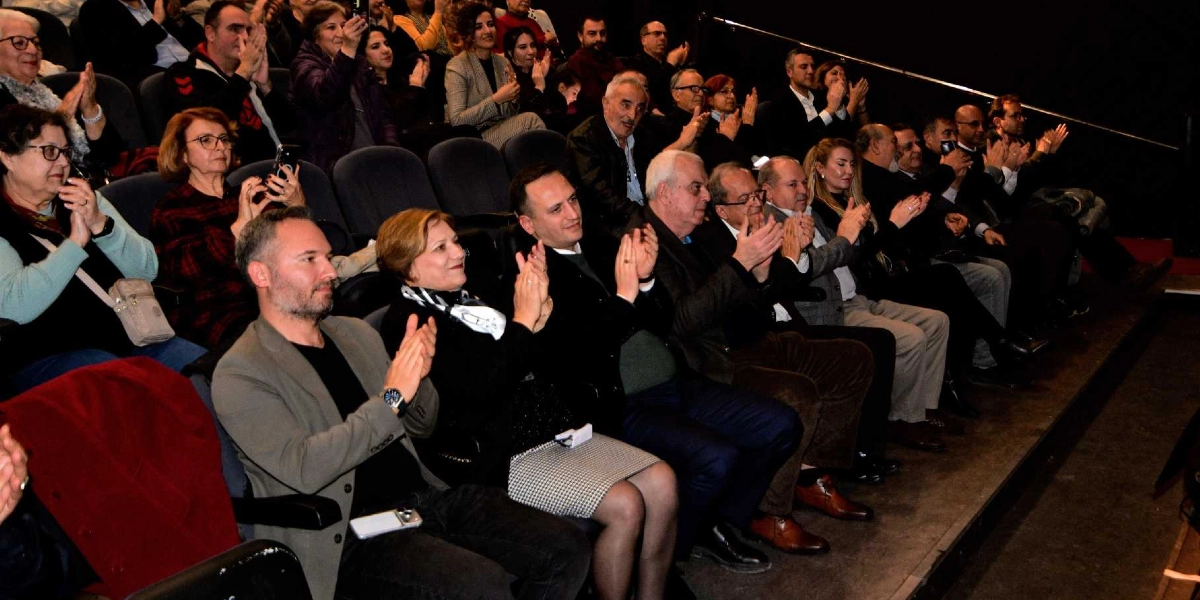 Kapalı bir salonda, tiyatro veya konferans koltuklarında oturan kalabalık bir izleyici grubu, sahnedeki bir etkinliği alkışlıyor. Ön sırada, gri ceketli bir erkek, siyah kıyafetli ve boynunda beyaz desenli bir fular olan bir kadın, yanında koyu renk ceketli bir erkek ve diğer katılımcılar yer alıyor. İzleyicilerin çoğu gülümseyerek veya dikkatle sahneye bakarak elleriyle alkış tutuyor, bazıları ise cep telefonlarıyla çekim yapıyor. Ortam loş bir ışıkla aydınlatılmış.
