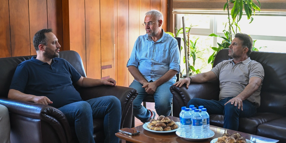 Koyu renkli deri koltuklarda oturan üç erkek, ahşap kaplamalı bir odada sohbet ediyor. Lacivert tişörtlü bir kişi, açık mavi gömlekli ve bej polo yaka tişörtlü iki kişiyle karşılıklı oturarak konuşuyor. Önlerindeki alçak masada bir tabak kurabiye ve birkaç şişe su bulunuyor.