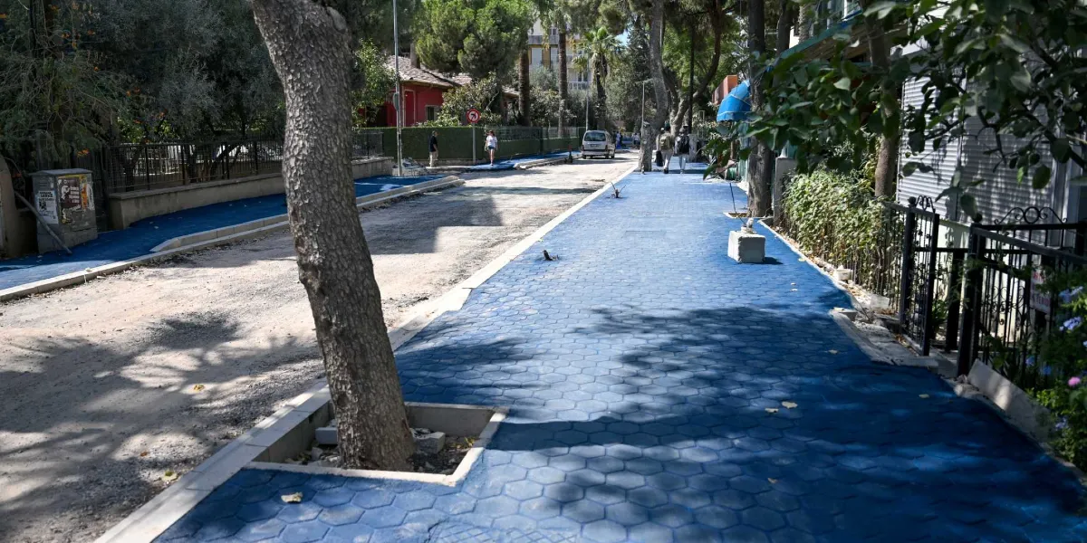 Yenilenmekte olan bir sokakta, her iki tarafı da parlak mavi renkli altıgen taşlarla döşenmiş geniş kaldırımlar ve ortada henüz asfaltlanmamış toprak bir yol bulunuyor. Kaldırımların kenarlarında ağaçlar ve binaların bahçe çitleri yer alırken, arka planda yürüyen insanlar ve park halindeki bir otomobil görülüyor.
