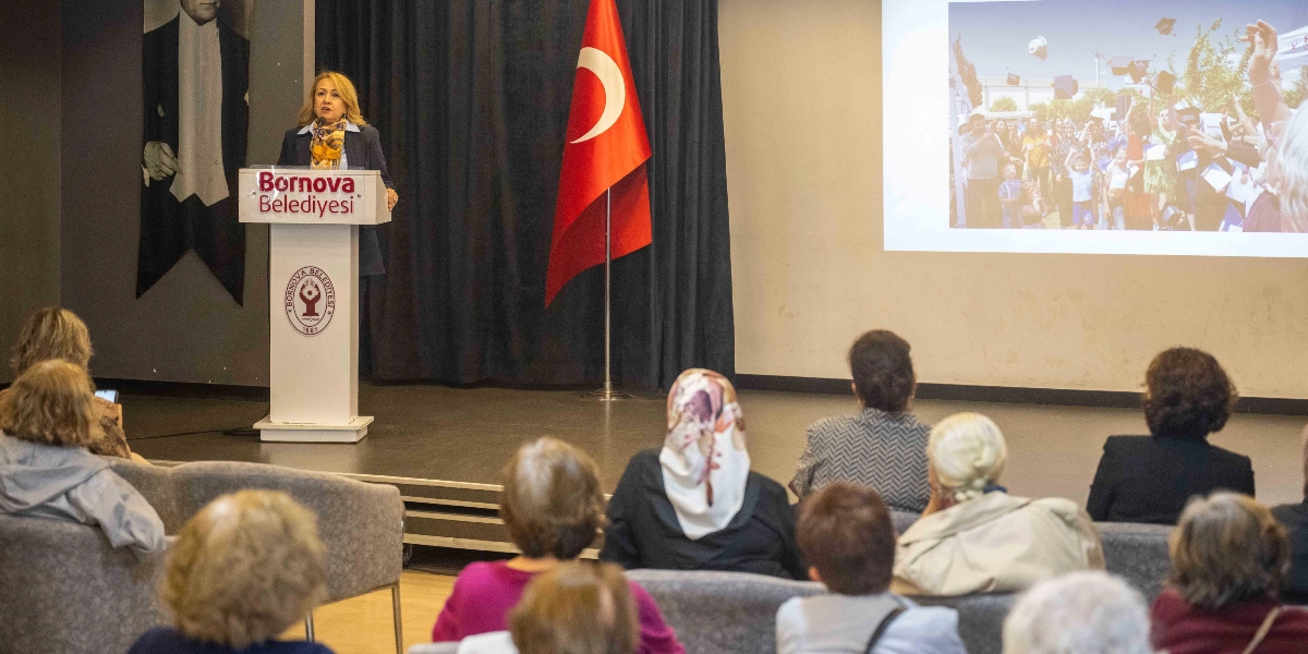 Bornova Belediyesi kürsüsünde konuşma yapan bir kadın, salondaki dinleyicilere hitap ediyor. Kürsünün arkasında Türk bayrağı ve Mustafa Kemal Atatürk’ün portresi yer alıyor. Sahnedeki dev ekranda ise mezuniyet kepi fırlatan mutlu bir grup insanın fotoğrafı yansıtılıyor. Salon, konuşmayı dinleyen katılımcılarla dolu.