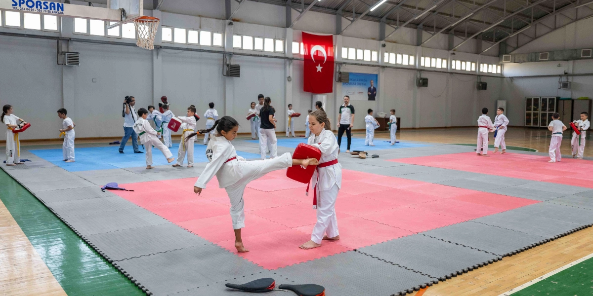 Kapalı bir spor salonunda tekvando antrenmanı yapan çocuklar; ön planda beyaz kıyafetli ve kırmızı kuşaklı iki kız öğrenci, birinin diğerinin tuttuğu kırmızı darbe yastığına tekme atmasıyla çalışıyor. Salonun zemininde pembe ve gri renkli minderler bulunuyor, arka planda ise Türk bayrağı asılı ve antrenman yapan diğer öğrenciler ile bir antrenör yer alıyor.