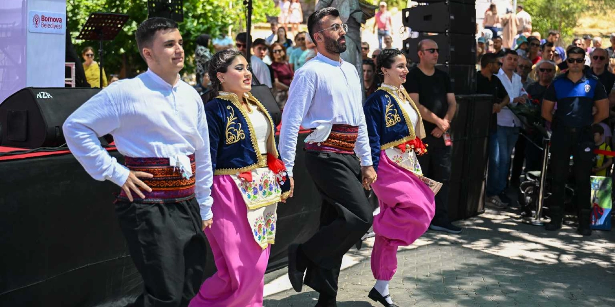 Geleneksel kıyafetler giymiş iki kadın ve iki erkekten oluşan bir grup, Bornova Belediyesi logolu bir sahnenin önünde halk oyunu sergiliyor. Erkekler beyaz gömlek, siyah pantolon ve desenli kuşak; kadınlar ise beyaz bluz, pembe şalvar, işlemeli lacivert yelek ve çiçek motifli önlük giyiyor. Arka planda etkinliği izleyen kalabalık bir grup ve ses sistemleri yer alıyor.