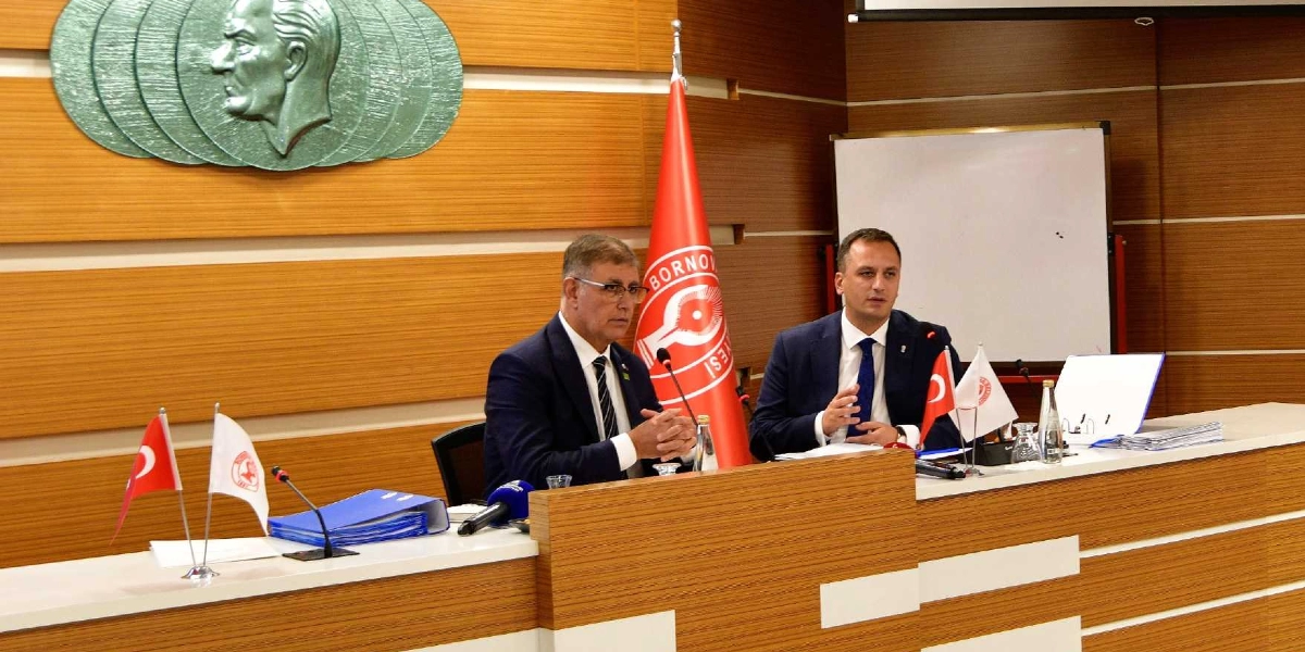 Ahşap kaplamalı bir toplantı salonunda, üzerinde Türk bayrağı ve Bornova Belediyesi'nin flaması bulunan bir masada oturan iki erkek yönetici, önlerindeki mikrofonlarla konuşma yapıyor. Arka planda duvarda Atatürk'ün kabartma silüeti ve Bornova Belediyesi'nin kırmızı renkli bayrağı yer alıyor.