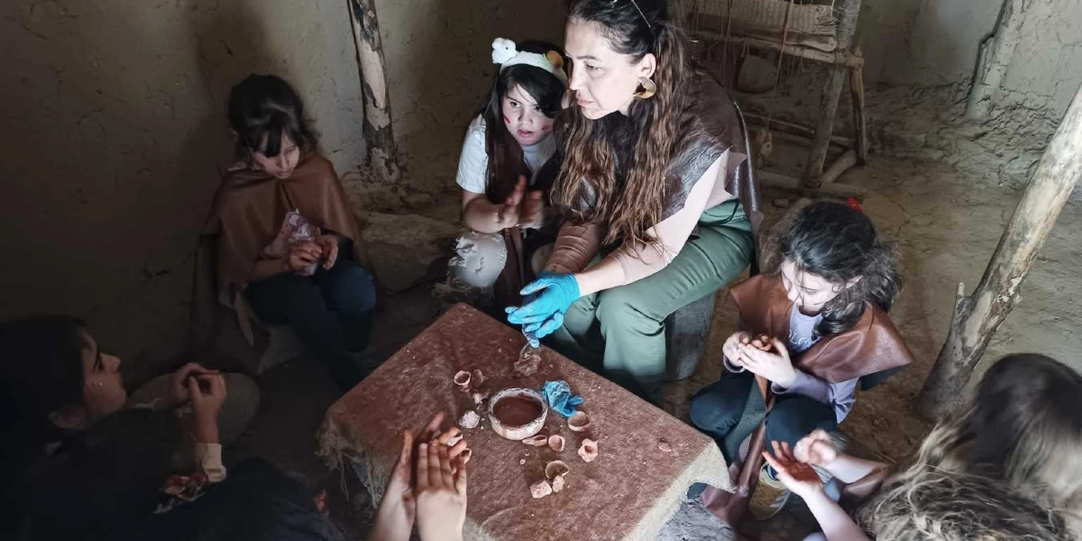 Taş Devri temalı bir atölye çalışmasında, üzerinde küçük çanak çömlek parçaları ve bir kap bulunan masanın etrafında toplanmış bir eğitmen ve beş çocuk. Eğitmen, ellerinde mavi eldivenlerle çocuklara çamurla çalışma konusunda rehberlik ediyor. Çocuklar, dönemi yansıtan kahverengi pelerinler giymiş ve çamurla uğraşarak Taş Devri yaşamını deneyimliyorlar. Ortam, kerpiç duvarları ve ahşap destekleriyle tarih öncesi bir kulübeyi andırıyor.