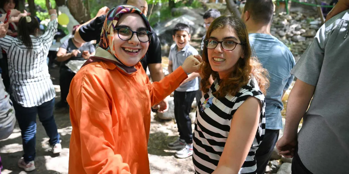 Açık havada, ağaçlık bir alanda bir araya gelmiş neşeli bir grup insan. Ön planda, turuncu bir gömlek ve renkli bir başörtüsü takan, gözlüklü genç bir kadın ile yanında siyah beyaz çizgili bir tişört giyen, yine gözlüklü başka bir genç kadın el ele tutuşarak gülümsüyor. Arka planda ise etkinliğe katılan diğer insanlar ve eğlenen çocuklar görülüyor.