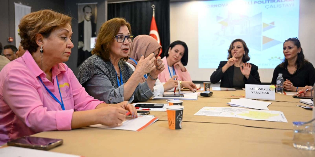 Bornova Erişilebilirlik Politikaları Çalıştayı'nda bir masa etrafında toplanmış, aralarında işaret diliyle iletişim kuran bir katılımcının da bulunduğu kadınlardan oluşan bir grup, toplantı masasında oturuyor. Masanın üzerinde 