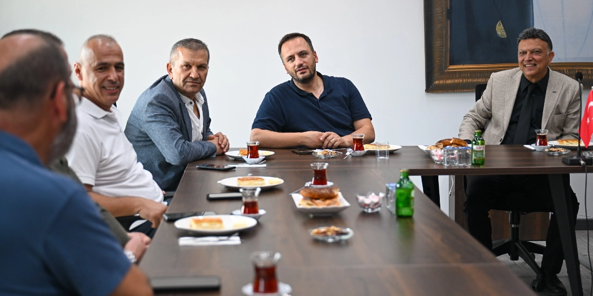 Beş erkek, üzerinde çay bardakları, tabaklar ve simitlerin bulunduğu uzun bir toplantı masasının etrafında oturuyor. Başkan Eşki, masanın orta kısmında lacivert bir tişörtle otururken, diğer katılımcılar günlük ve resmi kıyafetlerle ona eşlik ediyor. Masanın sağ ucunda, üzerinde Türk bayrağı bulunan bir masa bayrağı yer alıyor.