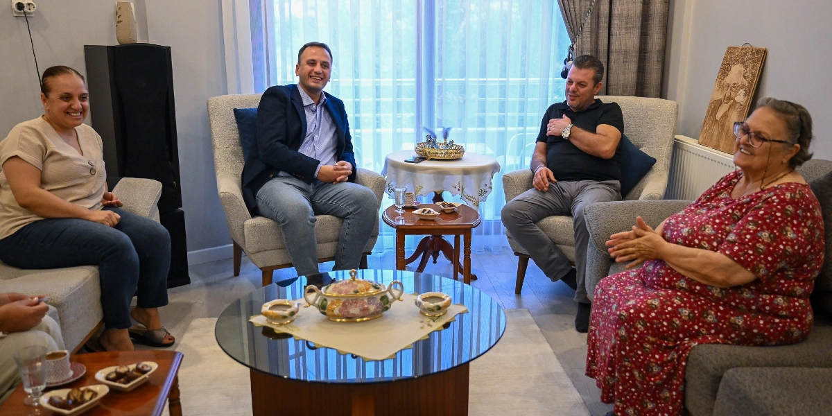 Bornova Belediye Başkanı Ömer Eşki, ev ortamında gerçekleşen bir ziyarette yaşlı bir kadın ve diğer misafirlerle birlikte oturup sohbet ediyor. Başkan Eşki, mavi gömlek ve lacivert ceket giymiş şekilde koltukta gülümseyerek otururken, karşısında çiçek desenli kırmızı bir elbise giyen yaşlı bir kadın ve yanında siyah tişörtlü bir erkek bulunuyor. Odanın ortasında üzerinde ikramlıkların olduğu cam bir sehpa, arka planda ise üzerinde Atatürk portresi bulunan bir tablo yer alıyor.