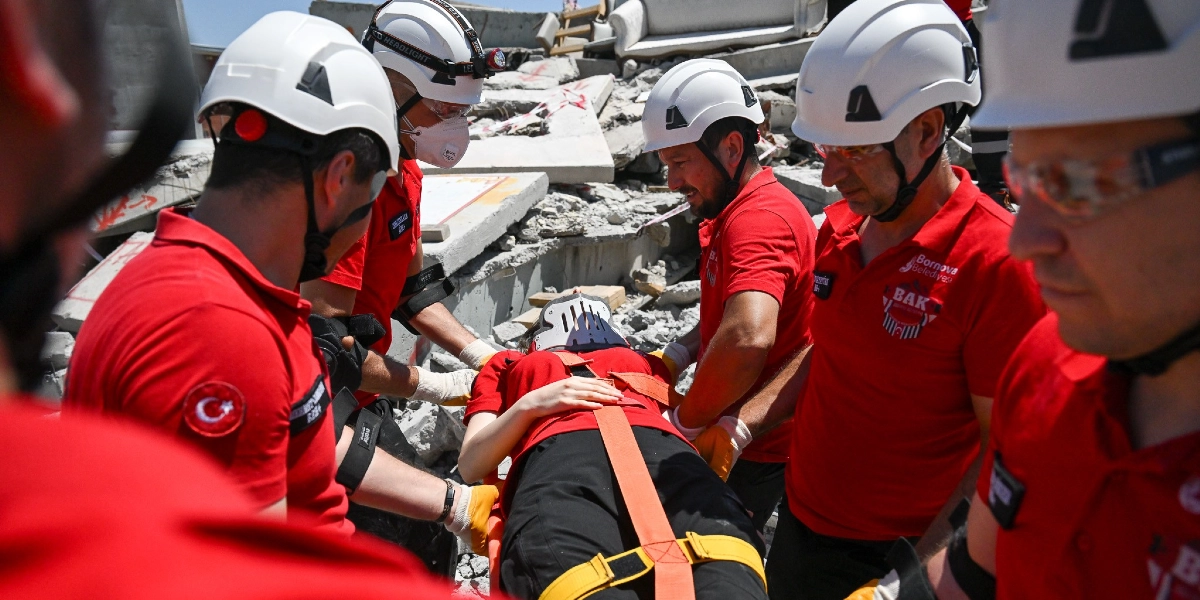 Beyaz baretli ve kırmızı üniformalı Bornova Belediyesi Arama Kurtarma ekibi, boyunluk takılmış yaralı bir kişiyi sedye üzerinde enkaz alanından dikkatle taşıyor.