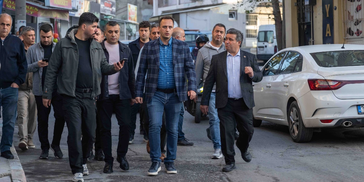 Bornova sokaklarında yürüyen, aralarında Bornova Belediye Başkanı Ömer Eşki'nin de bulunduğu bir grup erkek, incelemelerde bulunmak üzere ilerliyor. Ömer Eşki, kareli ceketi ve mavi gömleğiyle grubun ön sıralarında yer alırken, yanındaki ve arkasındaki diğer yetkililer ve çalışma arkadaşları ona eşlik ediyor. Sağ tarafta park halinde beyaz bir otomobilin bulunduğu sokakta, grup bir yandan yürüyüp bir yandan çevreyi gözlemliyor.