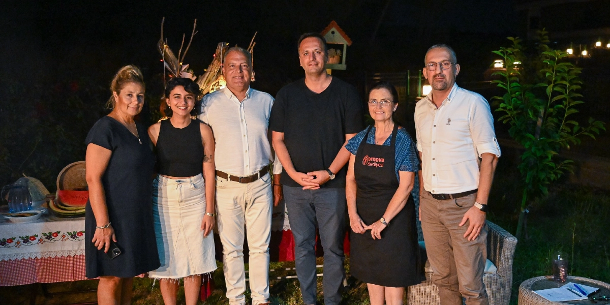 Gece vakti dış mekanda yan yana duran altı kişilik bir grup. Soldan sağa doğru; lacivert elbiseli bir kadın, siyah kolsuz bluz ve beyaz etek giyen genç bir kadın, açık renk gömlek ve pantolon giyen bir erkek, siyah tişörtlü bir erkek, üzerinde Bornova Belediyesi logosu bulunan siyah önlüklü bir kadın ve beyaz gömlekli bir erkek yer alıyor. Grubun sol tarafında, üzerinde karpuz dilimleri ve çeşitli yiyeceklerin olduğu, işlemeli bir örtüyle kaplı masa bulunuyor. Arka planda ise hafif aydınlatılmış bir bahçe ortamı görülüyor.