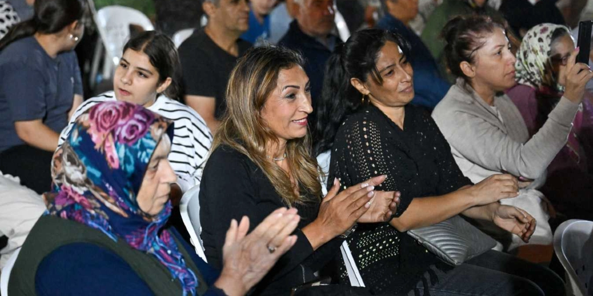 Açık havada düzenlenen bir etkinlikte yan yana oturan ve alkışlayarak etkinliğe katılan bir grup kadın izleyici. Ön planda başörtülü bir kadın, yanında siyah bluzlu ve uzun saçlı bir kadın ile siyah dantel detaylı bluz giyen diğer bir kadın yer alıyor. En sağda ise elindeki cep telefonuyla çekim yapan bir kadın görülüyor. İzleyicilerin arkasında ise etkinliği takip eden diğer katılımcılar bulunuyor.