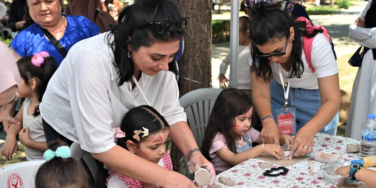Bornova Büyükpark'ta düzenlenen bir etkinlikte, beyaz tişörtlü iki kadın eğitmen, yüzleri boyalı çocuklara kil hamuruyla şekil verme konusunda yardımcı oluyor. Masanın üzerinde çeşitli kalıplar, kil parçaları ve bir şişe su bulunurken, eğitmenlerden birinin boynunda Bornova Belediyesi logolu 