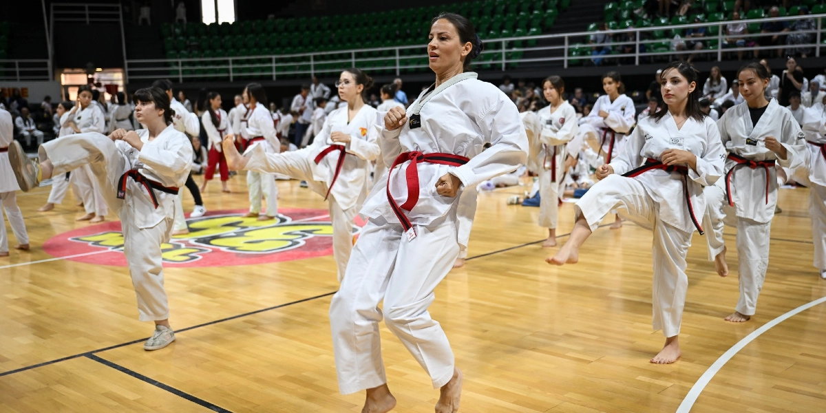 Kapalı bir spor salonunda, beyaz tekvando kıyafetleri ve kırmızı-siyah kuşaklar giymiş bir grup sporcu, eğitmenleri eşliğinde aynı anda tekme atma çalışması yapıyor. Ahşap zeminli sahada, ön planda odaklanmış bir şekilde hareketlerini sergileyen sporcuların arkasında, tribünlerde oturan izleyiciler ve diğer katılımcılar yer alıyor.