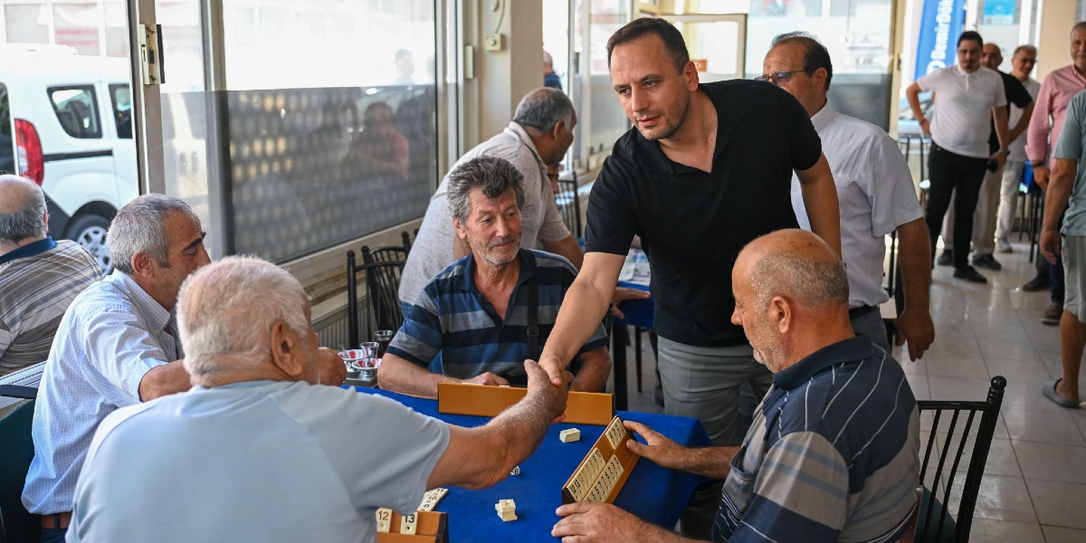 Siyah tişörtlü bir erkek, bir kıraathanede okey oynayan yaşlı bir adamla el sıkışıyor. Masada okey ıstakaları ve taşlar bulunurken, çevredeki diğer vatandaşlar oyunu izliyor.
