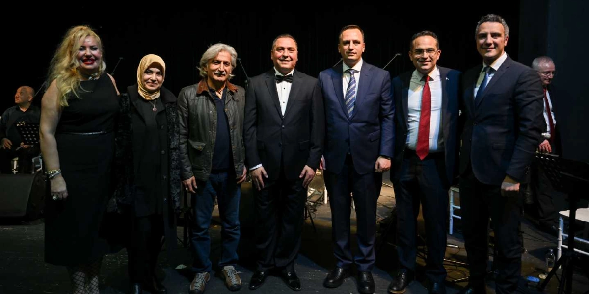 Sahne üzerinde yan yana duran, aralarında Bornova Belediye Başkanı Mustafa İduğ'un da bulunduğu yedi kişilik grup, bir konser veya etkinlik sonrası izleyicilere gülümseyerek poz veriyor. Grupta siyah elbiseli bir kadın, başörtülü bir kadın, deri ceketli bir erkek ve takım elbiseli dört erkek yer alıyor. Arka planda siyah bir sahne perdesi ve müzik ekipmanları görülüyor.