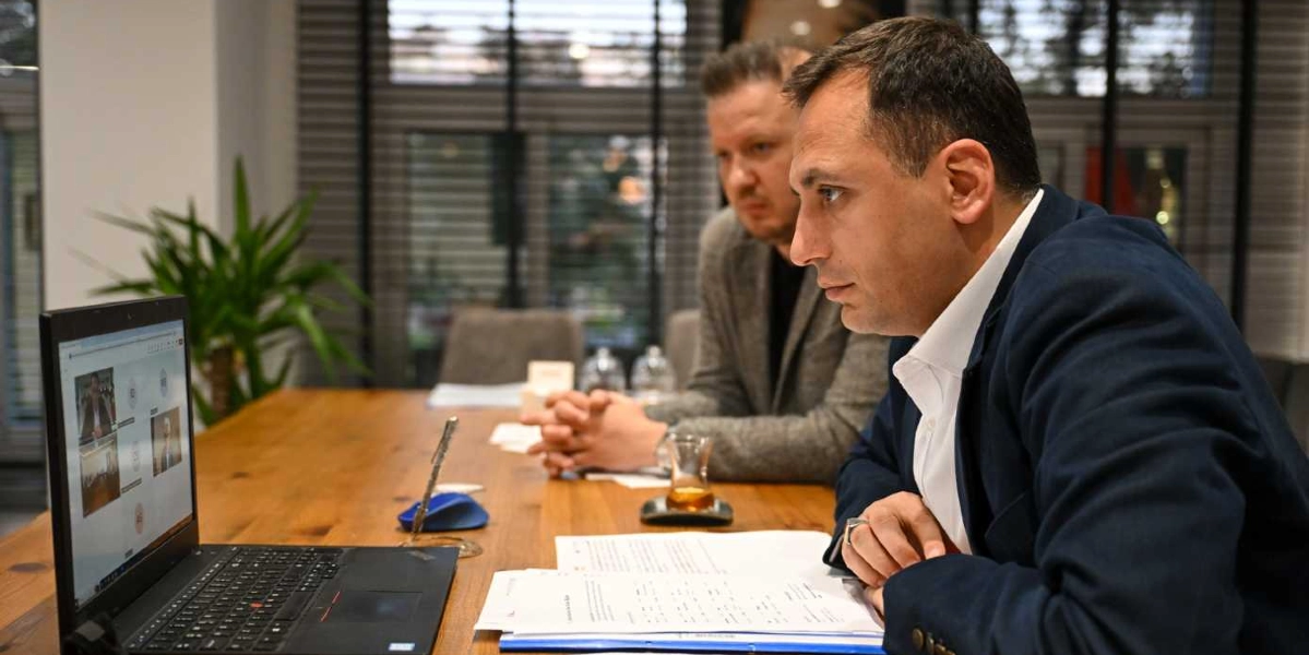Ofis ortamında ahşap bir masada oturan iki erkek, dizüstü bilgisayar ekranından çevrimiçi bir toplantıya katılıyor. Ön planda yer alan kişi lacivert ceketli, arka planda oturan kişi ise gri ceketlidir. Masanın üzerinde belgeler, bir kalem ve ince belli bir bardakta çay bulunmaktadır.