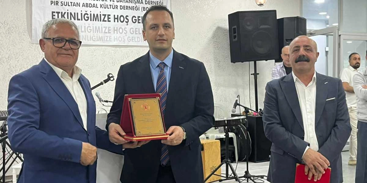 Kapalı bir mekanda, Pir Sultan Abdal Kültür Derneği Bornova Şubesi'nin düzenlediği etkinlikte, takım elbiseli üç erkek yan yana duruyor. Ortadaki kişi, kendisine takdim edilen plaketi iki eliyle tutuyor. Sol taraftaki kişi gözlüklü, sağ taraftaki kişi ise bıyıklı ve elinde kırmızı bir dosya tutuyor. Arka planda 