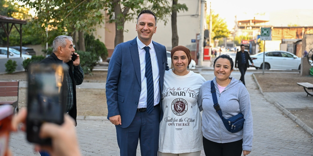 Mavi takım elbiseli ve kravatlı bir erkek, dış mekanda iki kadınla yan yana durarak gülümsüyor. Kadınlardan biri üzerinde İngilizce yazılar bulunan beyaz bir tişört, diğeri ise gri bir kapüşonlu üst ve çapraz çanta giyiyor. Sol tarafta telefonla konuşan yaşlı bir adam ve ön planda fotoğraf çeken birinin eli görülüyor. Arka planda ise ağaçlar, park bankları ve park halindeki araçların bulunduğu bir sokak yer alıyor.
