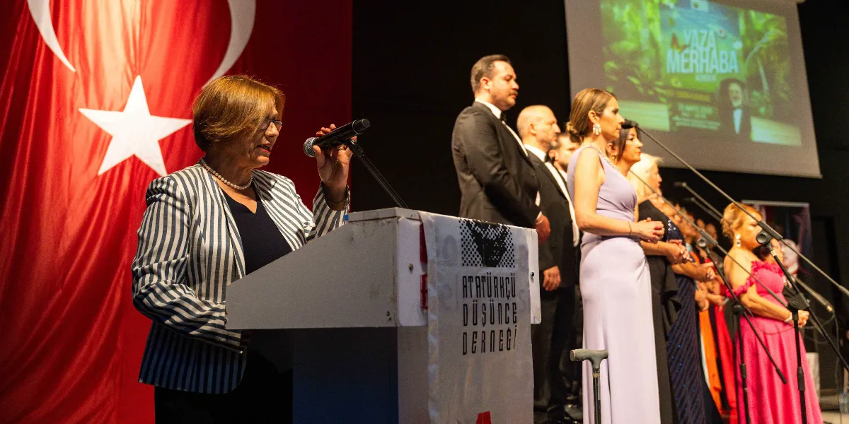 Kürsüde konuşma yapan gözlüklü, çizgili ceketli bir kadın ve arkasında Atatürkçü Düşünce Derneği yazılı kürsünün yanında duran, şık kıyafetler içindeki bir grup insan görülüyor. Arka planda büyük bir Türk bayrağı ve üzerinde 