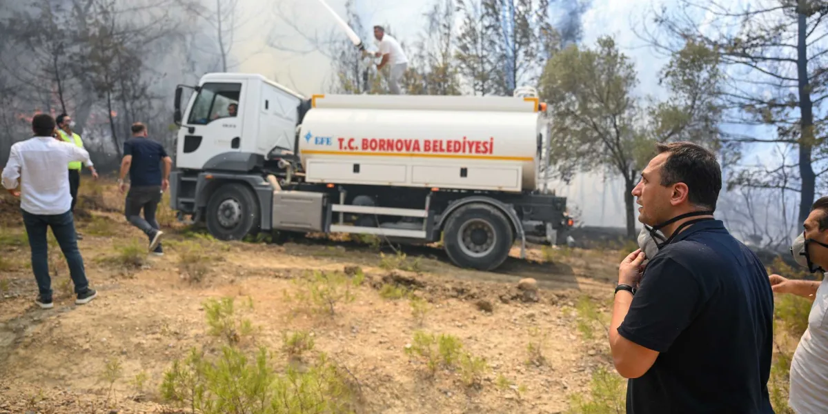 Gökdere'deki orman yangınına müdahale eden Bornova Belediyesi'ne ait bir arazöz, ağaçların arasından yükselen dumanların önünde duruyor. Aracın üzerinde bulunan bir görevli hortumla yangına su sıkarken, çevredeki belediye personeli ve yetkililer çalışmaları yakından takip ediyor. Ön planda, yüzünde maske bulunan bir yetkili endişeli bir ifadeyle yangın bölgesine bakıyor.