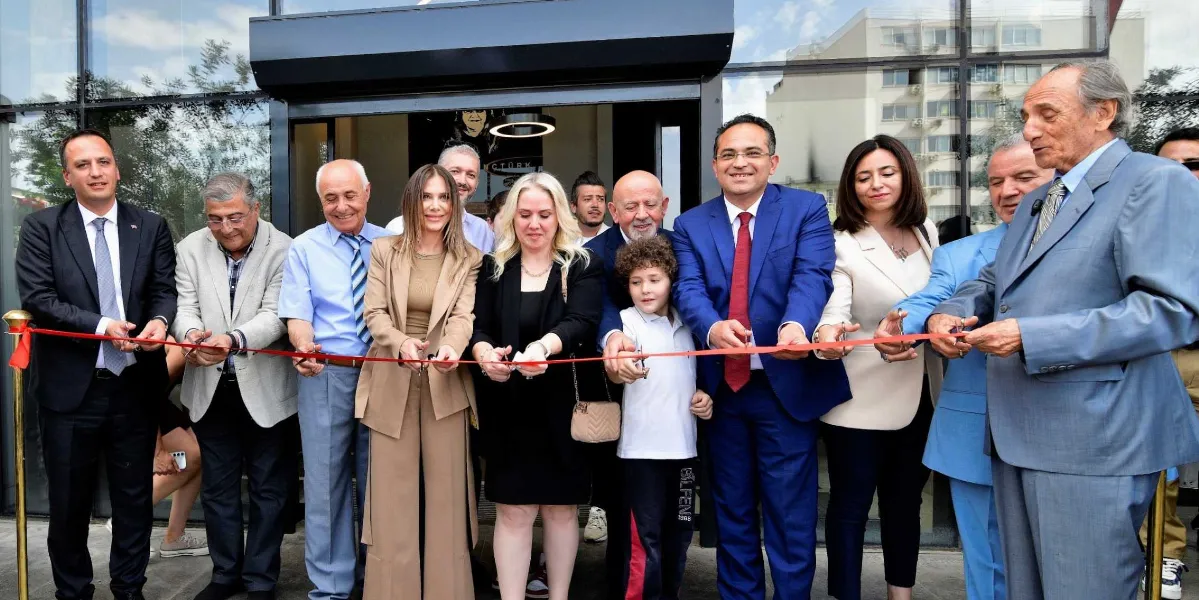 Bornova Belediyesi yetkilileri ve davetlilerin, bir binanın açılış töreni kapsamında kırmızı kurdeleyi hep birlikte kestiği an. Grupta takım elbiseli erkekler, şık giyimli kadınlar ve aralarında bir çocuğun da bulunduğu katılımcılar, modern bir cam binanın önünde yan yana dizilmiş şekilde ellerindeki makaslarla kurdeleyi tutuyor.