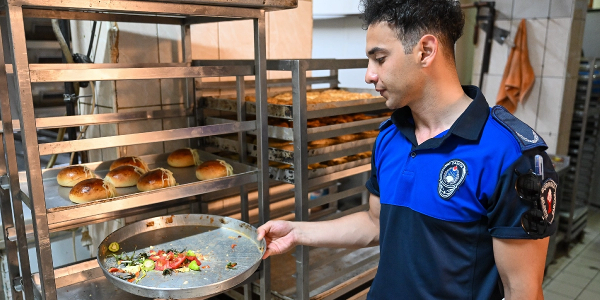 Bornova Belediyesi zabıta üniformalı bir görevli, bir fırın mutfağındaki metal rafın önünde durarak elinde tuttuğu tepsideki yemek artıklarını inceliyor. Yanındaki metal raflarda ise taze pişmiş poğaçalar dizili tepsiler bulunuyor.