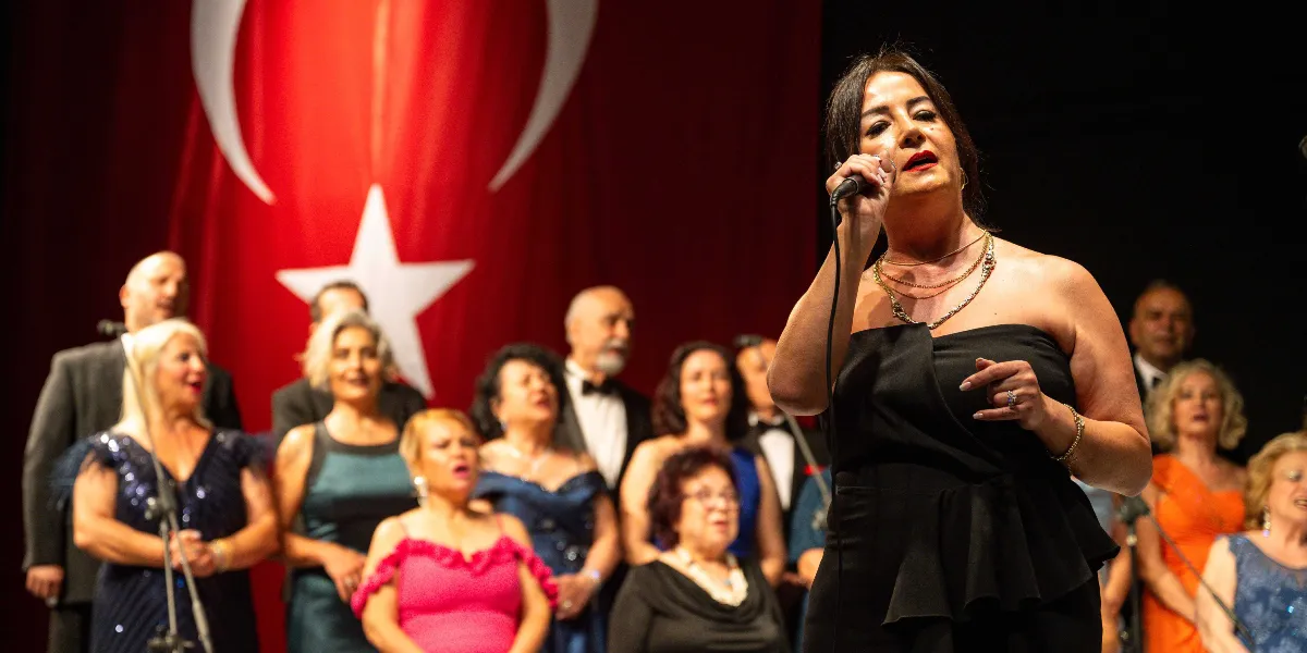 Siyah straplez elbise giymiş bir kadın solist, elinde mikrofonla sahnede şarkı söylüyor. Arka planda, üzerinde büyük bir Türk bayrağı asılı olan sahnede, şık kıyafetler içindeki koro üyeleri ona eşlik ediyor.