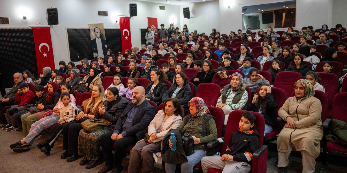 Konferans salonunda düzenlenen sertifika töreninde, kırmızı koltuklarda oturan çok sayıda genç ve aileleri sahneye doğru bakıyor. Salonun ön kısmında, duvarda asılı Türk bayrakları ve Mustafa Kemal Atatürk portresi yer alıyor. Katılımcılar arasında farklı yaş gruplarından öğrenciler ve yetişkinler bulunuyor.
