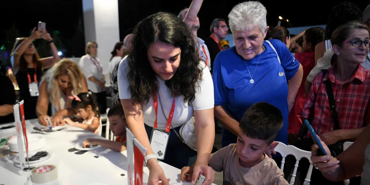 Uluslararası İzmir Fuarı'ndaki Bornova Belediyesi standında, beyaz bir masa etrafında toplanmış çocuklar ve yetişkinler el işi etkinlikleri yapıyor. Beyaz tişörtlü ve boynunda görevli kartı bulunan bir kadın, masadaki bir çocuğa yardımcı oluyor. Yanında mavi tişörtlü, kısa beyaz saçlı bir kadın gülümseyerek onları izliyor. Arka planda ise etkinliği takip eden veya fotoğraf çeken diğer ziyaretçiler bulunuyor.