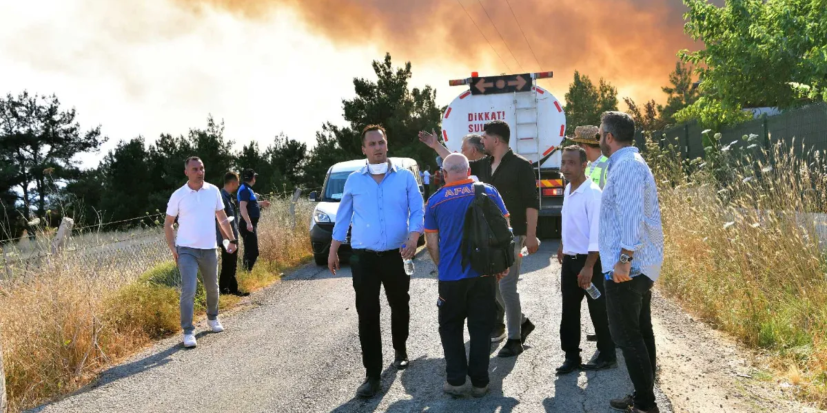 Yakaköy'deki orman yangını bölgesinde, asfalt bir yol üzerinde bulunan bir grup yetkili ve görevli, arka planda yükselen yoğun dumanlara doğru bakıyor. Grubun hemen arkasında üzerinde 