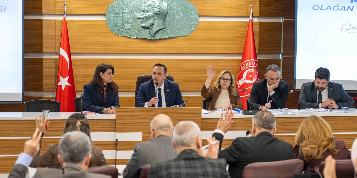 Bornova Belediyesi meclis salonunda, masada oturan beş kişilik bir heyet ve onları dinleyen katılımcıların yer aldığı bir toplantı. Masanın ortasında konuşma yapan bir yetkili, yanında ise diğer meclis üyeleri bulunuyor. Arka planda duvarda Atatürk kabartması, Türk bayrağı ve Bornova Belediyesi flaması yer alıyor. Toplantıya katılan bazı kişiler ellerini kaldırarak oylamaya veya görüş bildirmeye katılıyor.