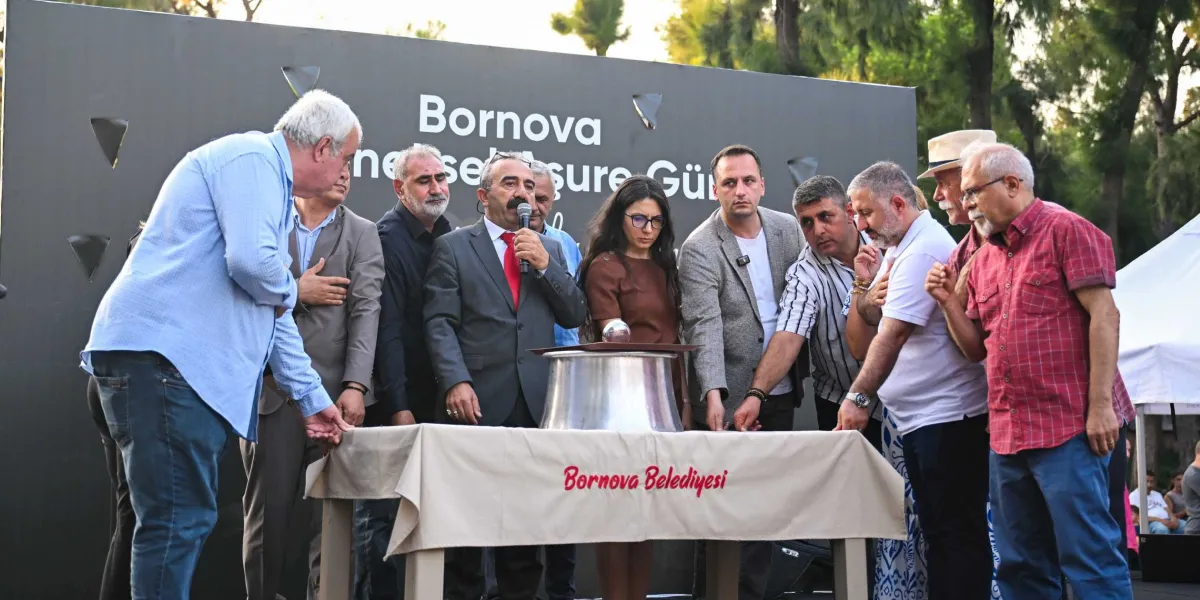 Bornova Belediyesi tarafından düzenlenen Geleneksel Aşure Günü etkinliğinde, üzerinde Bornova Belediyesi yazılı örtü serili bir masanın etrafında toplanmış bir grup insan yer alıyor. Ortada mikrofonla konuşma yapan takım elbiseli bir yetkili ve yanında duran diğer katılımcılar, masanın üzerindeki büyük metal kazana ellerini uzatarak aşure dağıtımını sembolize ediyorlar. Arka planda üzerinde Bornova Geleneksel Aşure Günü yazan büyük bir pano bulunuyor.