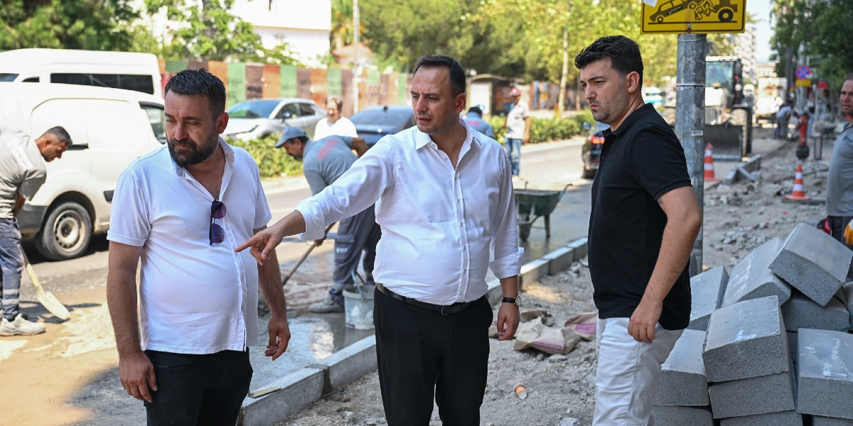 Beyaz gömlekli bir erkek, yanındaki kişiye eliyle yol çalışması yapılan alanı işaret ederek bir şeyler anlatıyor. Yanlarında siyah tişörtlü bir başka erkek onları dinliyor. Arka planda iş kıyafetleri giymiş işçiler, yol kenarında yığılı beton bloklar ve park halindeki araçların bulunduğu bir sokak çalışması görülüyor.