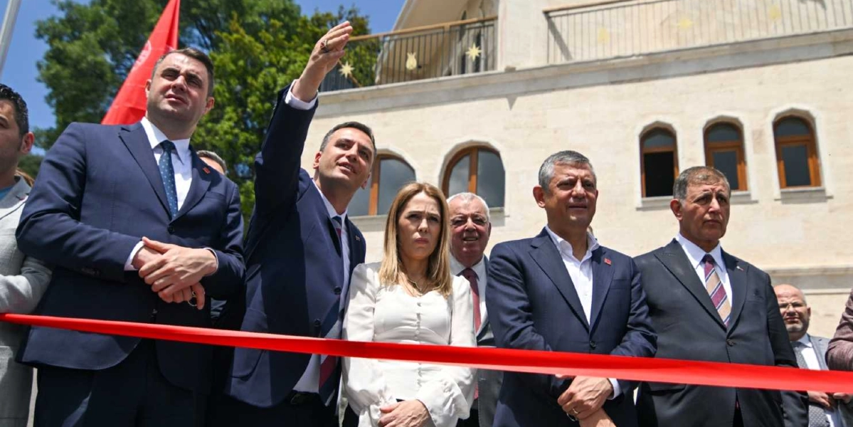CHP Genel Başkanı Özgür Özel ve beraberindeki heyet, Bornova'da bir açılış töreninde kırmızı kurdele önünde yan yana duruyor. Özgür Özel'in sağ tarafında bulunan bir yetkili eliyle ileriyi işaret ederken, grup önlerindeki kırmızı kurdelenin kesilmesini bekliyor. Arka planda taş mimariye sahip bir bina ve ağaçlar görülüyor.