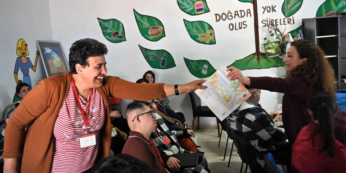 Kırmızı beyaz çizgili tişört ve kahverengi hırka giyen gülen bir kadın, elindeki bir kitabı karşısındaki kişiye uzatıyor. Arka planda duvarda, atıkların doğada yok olma sürelerini gösteren yaprak şeklinde çizimlerin olduğu bir ağaç figürü yer alıyor. Sınıf ortamındaki etkinlikte, özel bireyler ve eğitmenler bir arada bulunuyor.