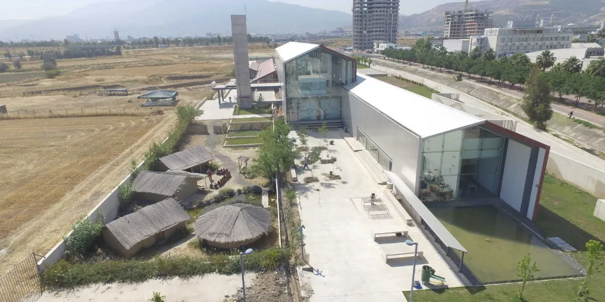 Bornova'da yer alan modern mimariye sahip, cam cepheli ve uzun çatılı müze binası ile hemen yanındaki geleneksel sazdan yapılmış kulübeleri içeren açık hava sergileme alanı. Binanın önünde geniş bir yürüyüş yolu, küçük bir süs havuzu ve çevre düzenlemesi bulunurken, arka planda inşaat halindeki yüksek yapılar ve geniş bir arazi görülüyor.