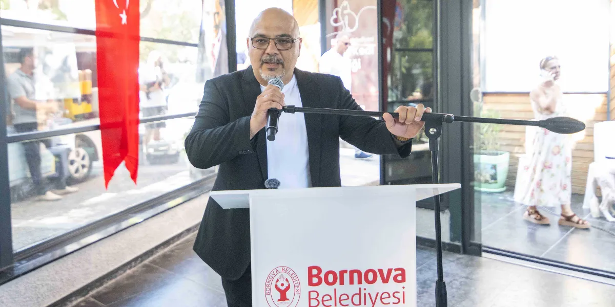 Kürsüde konuşma yapan, gözlüklü, kirli sakallı ve siyah ceketli bir erkek. Önündeki beyaz kürsü üzerinde Bornova Belediyesi logosu ve yazısı yer alıyor. Arka planda cam bölmenin ardında Türk bayrağı, ayakta duran insanlar ve dış mekan görülüyor.