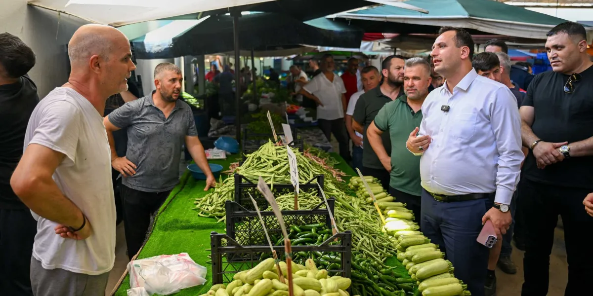 Bornova Belediye Başkanı Ömer Eşki, bir pazaryerindeki tezgahları ziyaret ederek esnafla sohbet ediyor. Başkan Eşki, beyaz gömlek ve lacivert pantolon giymiş şekilde, önündeki taze sebzelerle dolu tezgahın başında duran esnaf ve çevresindeki vatandaşlarla konuşuyor. Tezgahın üzerinde yeşil fasulye, kabak ve biber gibi ürünler sergileniyor.