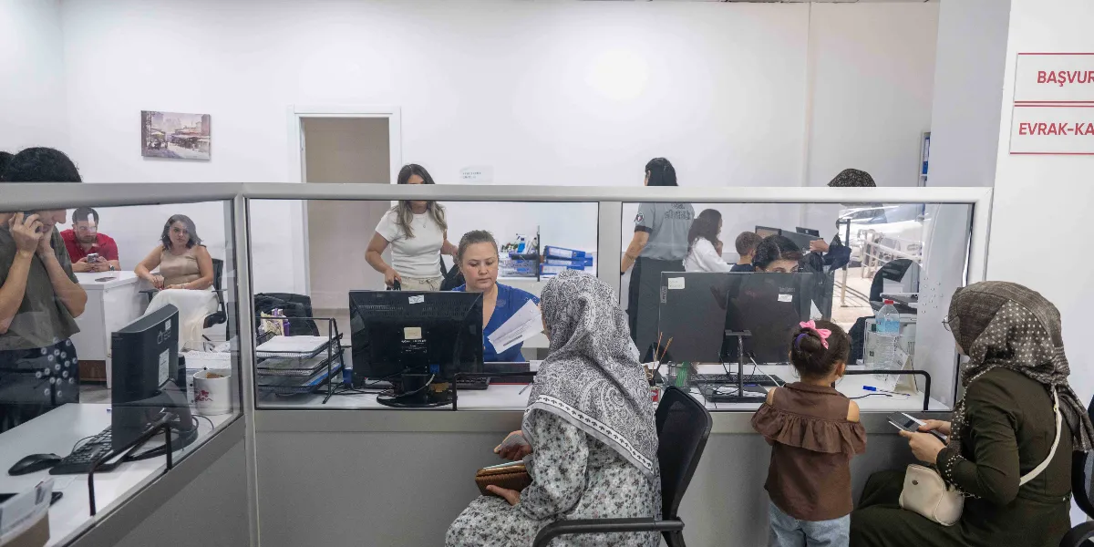 Modern bir ofis ortamında, cam bölmelerle ayrılmış bankolarda çalışan görevliler ve işlem yapmak için bekleyen vatandaşlar bulunuyor. Ön planda, başörtülü bir kadın ve yanında küçük bir kız çocuğu, görevliyle görüşmek için bekliyor. Arka planda ise masalarında bilgisayarlarıyla çalışan diğer personel ve ofis içinde hareket eden insanlar görülüyor. Duvarın sağ üst köşesinde kırmızı harflerle 