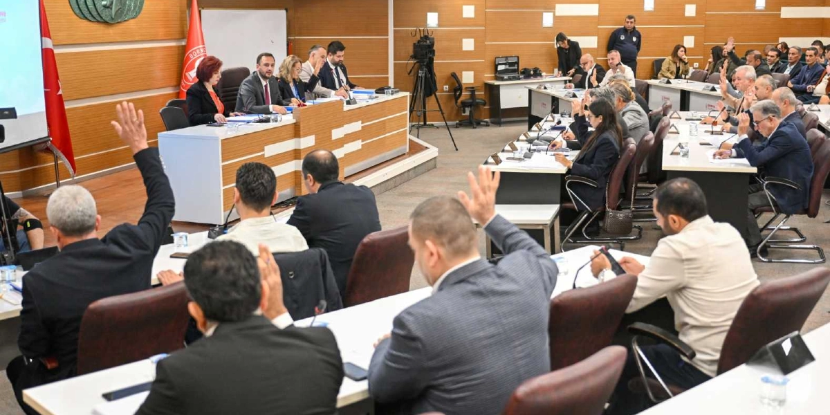 Bornova Belediyesi meclis salonunda düzenlenen toplantıda, katılımcıların büyük çoğunluğu ellerini kaldırarak oylama yapıyor. Salonun ön kısmında başkanlık divanı yer alırken, diğer üyeler U düzenindeki masalarda oturuyor. Arka planda Türk bayrağı ve Bornova Belediyesi'nin amblemi bulunuyor.
