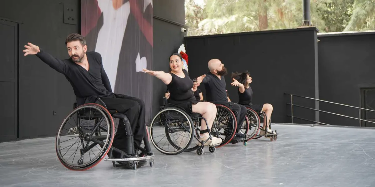 Siyah kıyafetler giymiş, tekerlekli sandalyedeki dört dansçı, açık hava sahnesinde kollarını yanlara açarak senkronize bir şekilde dans ediyor. Önde sakallı bir erkek ve yanında gülümseyen bir kadın, arkalarında ise diğer dansçılar yer alıyor. Arka planda siyah bir duvar ve üzerinde kısmen görünen bir portre bulunuyor.