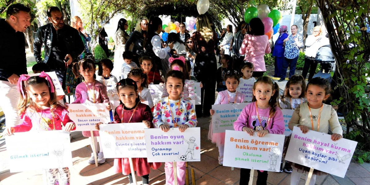 Açık havada, yeşillikler arasında düzenlenen Çocuk Hakları Günü etkinliğinde ellerinde çeşitli haklarını belirten dövizler tutan bir grup çocuk ve onlara eşlik eden yetişkinler yer alıyor. Ön sırada yan yana dizilmiş çocuklar, üzerinde 