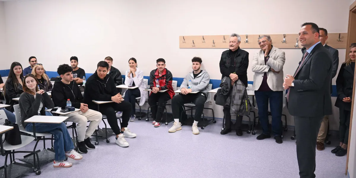 Bornova Belediye Başkanı Ömer Eşki, bir sınıfta toplanmış üniversite öğrencilerine hitap ediyor. Başkan Eşki, lacivert takım elbiseli ve kravatlı bir şekilde ayakta konuşurken, öğrenciler sıralarında oturmuş onu dinliyor. Sınıfta Başkan Eşki'nin yanında iki erkek misafir daha bulunuyor ve öğrenciler ilgiyle süreci takip ediyor.
