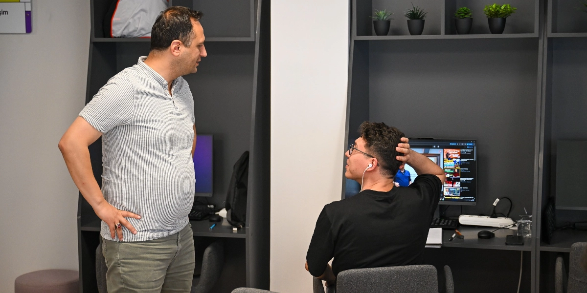 Modern bir ofis ortamında, gri çizgili tişört giyen bir erkek ayakta durarak, bilgisayar başında oturan ve kulaklıkla müzik dinleyen siyah tişörtlü başka bir erkekle sohbet ediyor. Bilgisayar başında oturan kişinin ekranında video içerikleri yer alıyor, arka planda ise raflarda küçük saksı bitkileri bulunuyor.